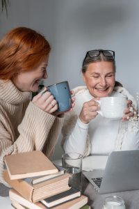 Two women laughing over coffee with books and laptop.