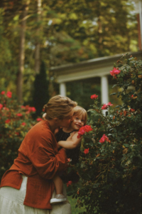 Woman holding child near rose bush in garden.